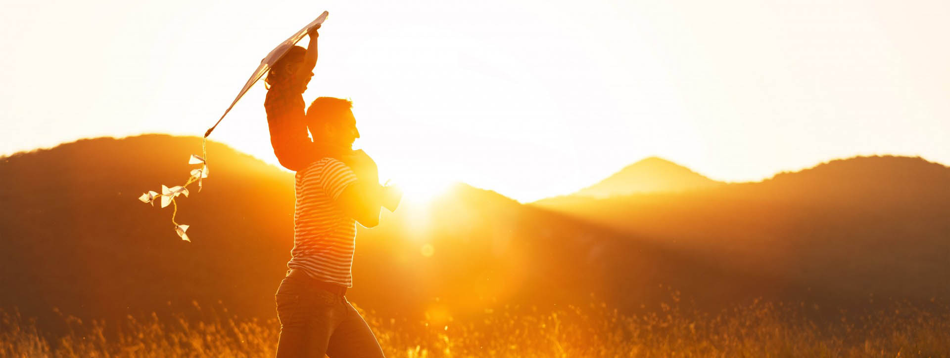 Dad with child on shoulders flying a kite during sunset